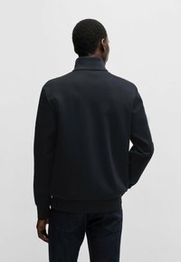 Black zip-up jacket with a high collar, featuring a smooth texture and ribbed cuffs. Paired with dark denim jeans.