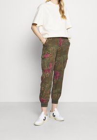 Olijfgroene cargobroek met een bloemenpatroon in roze en kastanjebruin, met zijzakken en elastische boorden. Gecombineerd met een witte top met korte mouwen en sneakers.