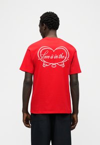 Άνδρας που φοράει κόκκινο T-shirt με λευκό σχέδιο σε σχήμα καρδιάς και κείμενο με καλλιγραφικά γράμματα «Love is in the» στην πλάτη, συνδυασμένο με μαύρο σορτς.