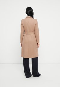 Frau mit langen schwarzen Haaren, die einen beigen Mantel mit Gürtel und weiten schwarzen Hosen trägt, steht vor einer schlichten weißen Wand.