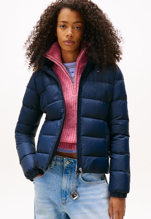 Ung kvinde med krøllet hår iført en mørkeblå dunjakke over en lyserød striktrøje og lyseblå jeans med en nøglering formet som et hjerte.