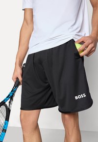 Črne športne kratke hlače z gladko teksturo, stranskim žepom, v katerem je teniška žogica, in belim napisom "BOSS" na spodnjem delu noge.