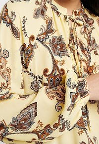 Yellow blouse made of lightweight fabric featuring a paisley pattern in brown, orange, and gray. Has a tie-neck detail and elasticized sleeves.