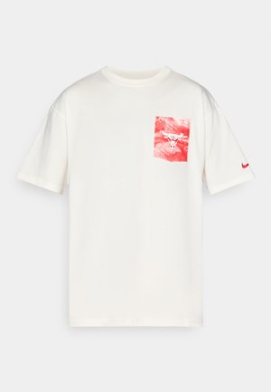 Kreemikotsane T-särk, millel on punase ja valge graafikuga tasku, millel on härja logo, ning väikese punase Nike swooshiga vasakul varrukal.