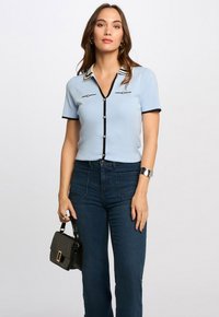 Hellbluse mit kurzem Ärmel und V-Ausschnitt, schwarzen Akzenten und perlenartigen Knöpfen, kombiniert mit dunklen Jeans und einer kleinen schwarzen Handtasche.