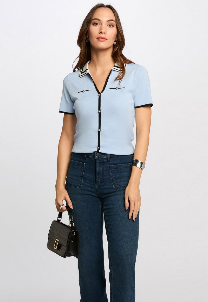 Hellbluse mit kurzem Ärmel und V-Ausschnitt, schwarzen Akzenten und perlenartigen Knöpfen, kombiniert mit dunklen Jeans und einer kleinen schwarzen Handtasche.