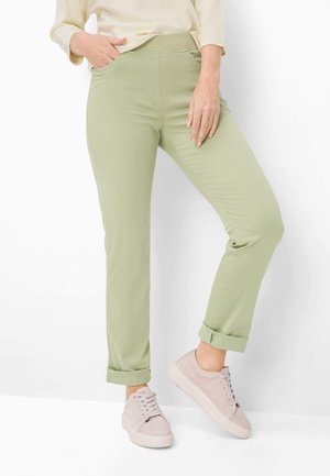 Frau trägt hellgrüne, hochgekrempelte Hose, blassrosa Schnürsneaker und ein cremefarbenes Langarmshirt, steht vor weißem Hintergrund.