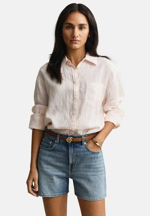 Frau mit langen dunklen Haaren, die ein hellrosa Hemd mit hochgekrempelten Ärmeln, Jeansshorts, braunen Gürtel und goldenes Armband trägt.