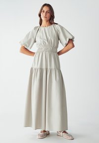 Robe maxi gris clair avec manches bouffantes et taille froncée. Présente une jupe à volants et un tissu doux. Portée avec des chaussures blanches en treillis.