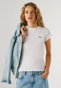 Noor naine blondide lokkis juustega, kannab valget kehakuju järgivat T-särki ja helesiniseid teksaseid, hoiab denimitagi üle õla.