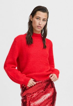 Pull rouge en tricot avec un col montant et des manches longues. Le tissu a l'air doux et moelleux. Associé à une jupe en sequins rouges brillants.