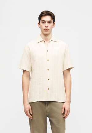 Junger Mann, der ein beiges, kurzärmeliges Hemd mit Knöpfen und olivgrüne Hosen trägt, vor einem schlichten hellen Hintergrund stehend.