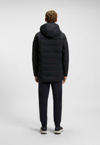 Black padded jacket with a hood, featuring quilted texture and a straight cut. Worn with dark pants and sneakers. Minimalist design.