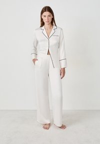 Femme debout pieds nus portant un ensemble de pyjama en satin crème avec passepoil noir, une main dans la poche contre un fond clair uni.