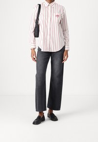 Camicia a righe con righe verticali rosse e bianche, abbinata a jeans a gamba larga grigio scuro e mocassini neri. Include una borsa a tracolla.