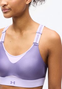 Κοντινό πλάνο ενός ατόμου που φοράει αθλητικό σουτιέν Under Armour σε λιλά απόχρωση με ρυθμιζόμενους ιμάντες με λογότυπο, πάνω σε απλό φόντο.