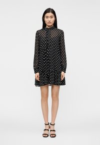 Νεαρή γυναίκα όρθια, φορώντας ένα μακρυμάνικο μαύρο διάφανο φόρεμα με λευκές πουά και μαύρα σανδάλια με ψηλό τακούνι, σε απλό λευκό φόντο.