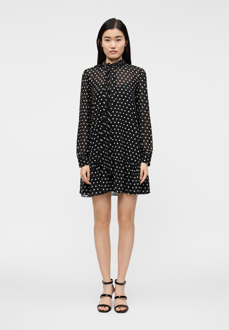 Νεαρή γυναίκα όρθια, φορώντας ένα μακρυμάνικο μαύρο διάφανο φόρε�μα με λευκές πουά και μαύρα σανδάλια με ψηλό τακούνι, σε απλό λευκό φόντο.