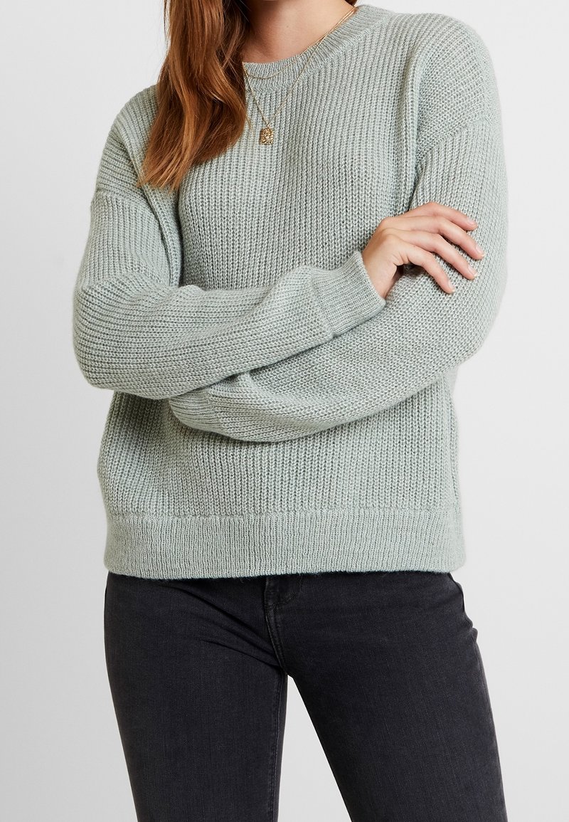 Femme portant un pull en laine gris clair et un jean noir, les bras croisés, avec des cheveux roux mi-longs et un collier pendentif en or.