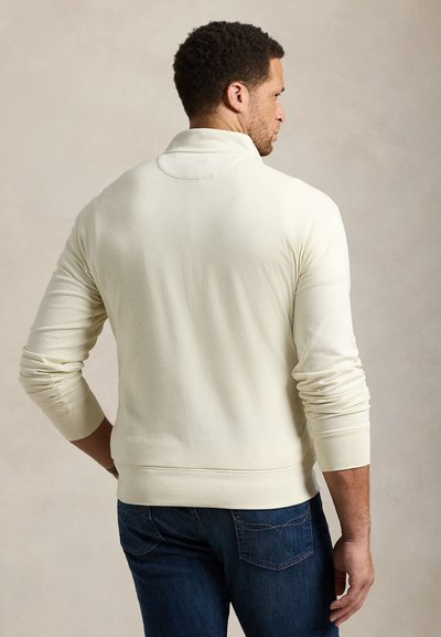 Ανοιχτό μπεζ πουλόβερ από μαλακό ύφασμα, με ψηλό γιακά και μακριά μανίκια, με ριμπ μανσέτες και τελείωμα, συνδυασμένο με σκούρο τζιν παντελόνι.