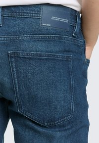 Tył osoby noszącej ciemnoniebieskie dżinsy z prostokątną skórzaną łatką z detalami marki. Ręka w bocznej kieszeni.