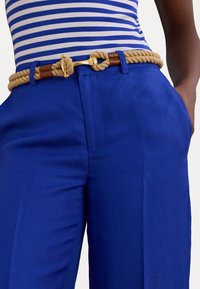 Pantalons larges bleus avec une texture lisse, ornés d'une ceinture en corde beige nouée et de détails en cuir marron. Top rayé partiellement visible.