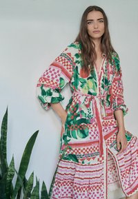 Rochie colorată cu motive florale și geometrice, mâneci lungi, curea de legare și fustă în volane, realizată dintr-un material ușor.