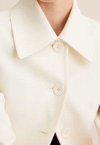 Crème-kleurige, gestructureerde blazer met een brede kraag, twee knopen en een gladde textuur; heeft minimale stiksels en een op maat gemaakte uitstraling.