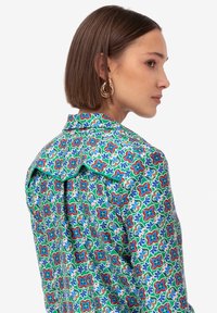 Camicia floreale colorata con una base verde vivace e motivi rossi e blu, colletto strutturato e tessuto leggero con una consistenza liscia.