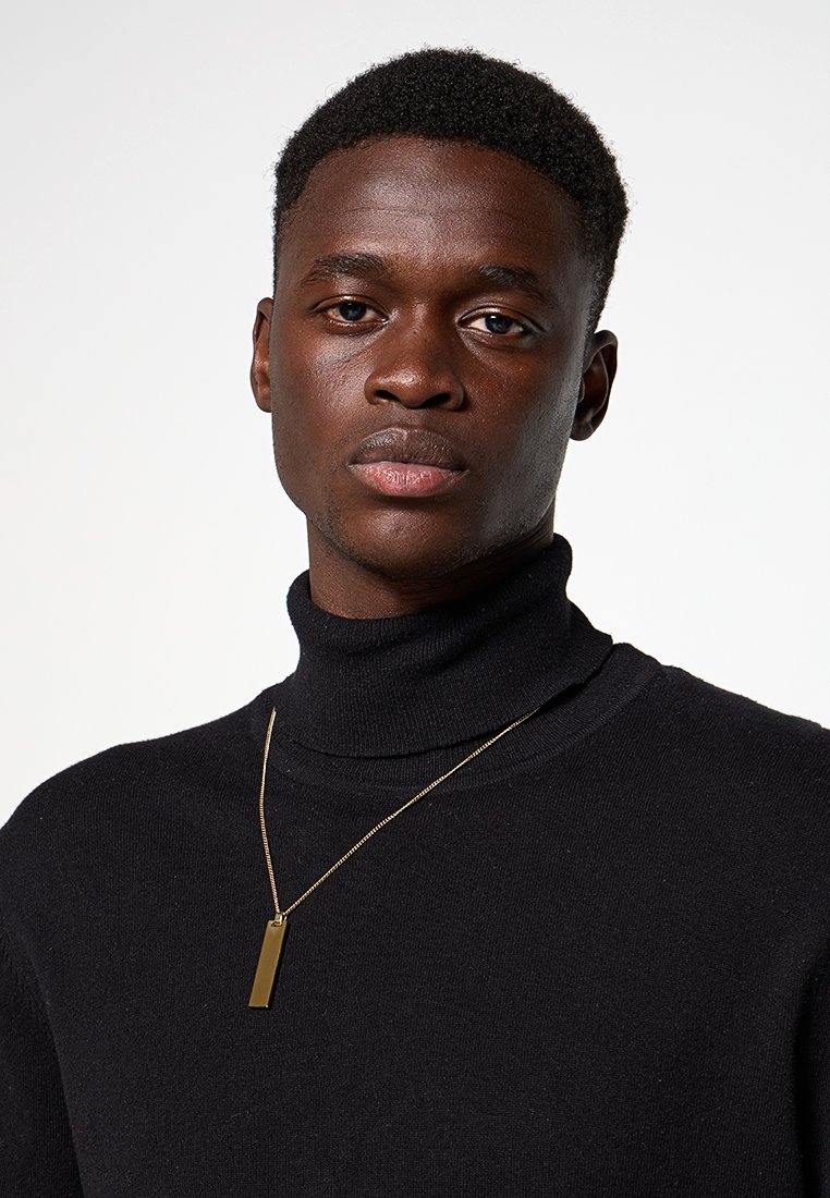 Junger schwarzer Mann mit kurzen lockigen Haaren, der einen schwarzen Rollkragenpullover und eine goldene rechteckige Anhängerkette trägt.