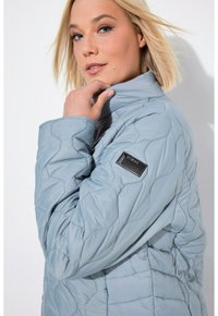 Chaqueta acolchada azul claro con un alto cuello y un patrón texturizado. Presenta una etiqueta de tela negra en la manga izquierda. Material suave.