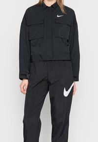 Svart Nike-jacka med två framfickor och en kortare design, kombinerad med matchande svarta byxor med en vit Nike-logga.