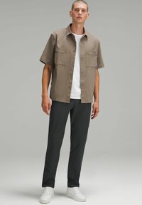 Chemise à manches courtes de couleur marron clair, avec deux poches avant et un design à boutons. Assortie à un pantalon sombre et des baskets blanches.