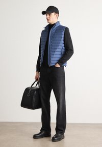 Chaleco acolchado azul claro, suéter negro de cuello alto, jeans oscuros y botas negras. Sosteniendo un bolso negro, usando una gorra negra. Estilo minimalista.