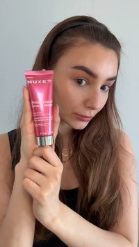 Vrouw met lang bruin haar houdt een roze NUXE Prodigieuse Hyaluronic Boost huidverzorgingstube dicht bij haar gezicht tegen een effen achtergrond.