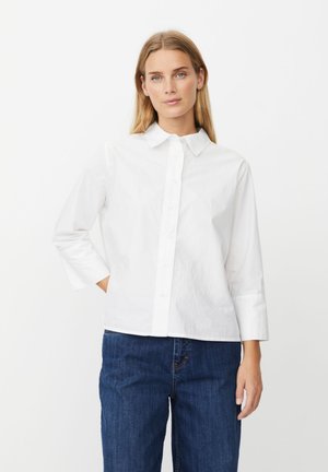 Kvinde med blondt hår iført en hvid skjorte med knapper og blå jeans, stående mod en ensartet lys baggrund, hånd i lommen.