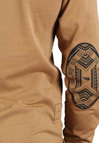 Chemise à manches longues en coton brun clair, présentant un motif tribal noir marquant au niveau des coudes et un détail d'étiquette sur le côté.