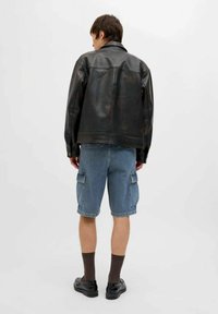 Giubbotto in pelle nera, pantaloni cargo in denim, scarpe nere, calze grigie. Modello mostrato di spalle su uno sfondo neutro.