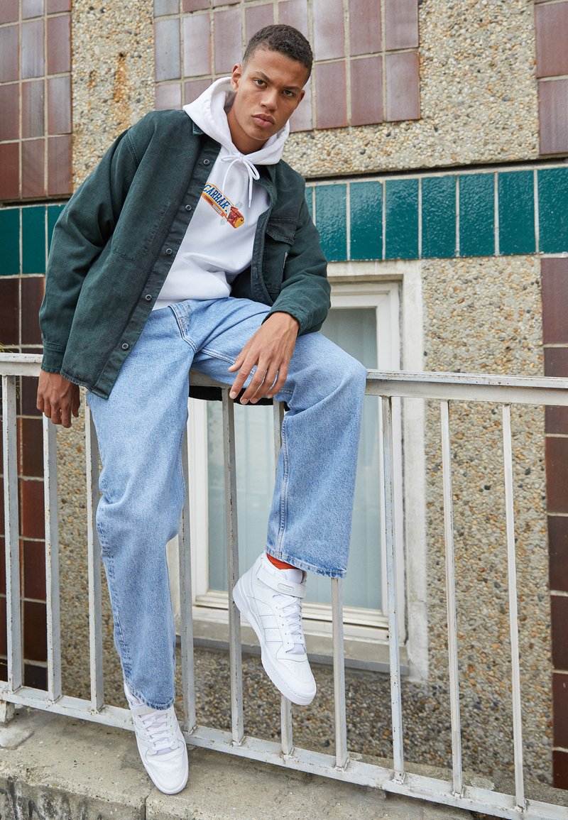 Model wearing a dark green button-up shirt over a white hoodie, light blue jeans, and white sneakers with orange accents, seated on a railing.