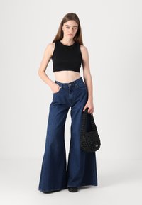 Musta crop-top, korkeavyötäröiset tummansiniset leveälahkeiset farkut, teksturoitu musta käsilaukku, korvissa renkaat, seisoo neutraalilla taustalla.