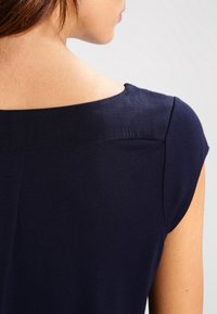 Detailní záběr na horní část zad a rameno osoby, která má na sobě námořnickomodrý top bez rukávů s širokým výstřihem.