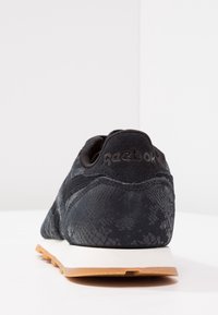 Svart Reebok sneaker med texturerat mönster, mocka hälla, vit mellansula och gummiyttersula, visad bakifrån på en vit yta.