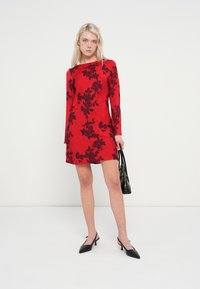Rdeča obleka z dolgimi rokavi in črnim cvetličnim vzorcem, dolžina nad koleni, gladke teksture, dopolnjena s črnimi čevlji z ostrim prstom in torbico.