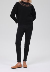 Czarny sweter dzianinowy z koronkowym wykończeniem przy dekolcie, zestawiony z czarnymi spodniami slim fit oraz beżowymi zamszowymi loafersami z metalicznymi akcentami.