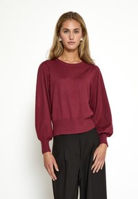 Burgunderfarbener Strickpullover mit Rundhalsausschnitt, puffigen Ärmeln und geripptem Saum. Kombiniert mit schwarzen, hoch taillierten Hosen.