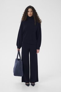 Γυναίκα με σγουρά μαλλιά που φοράει μαύρο ζιβάγκο, παντελόνι φαρδύ, μυτερά παπούτσια, κρατώντας μια μεγάλη τσάντα ώμου σκούρου μπλε, στέκεται σε λευκό φόντο.