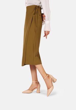 Gonna midi color khaki con allacciatura laterale, tessuto morbido e texture liscia; abbinata a scarpe con tacco a blocco beige e cinturino alla caviglia.