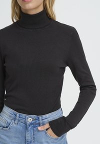 Pull noir à col roulé en maille côtelée, avec des manches longues aux poignets ajustés, associé à un jean en denim bleu clair.