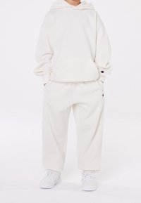 Barn i en överdimensionerad krämfärgad hoodie och matchande sweatpants med vita sneakers, händerna i fickorna mot en enkel ljus bakgrund.