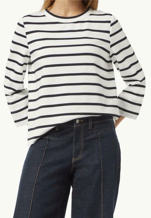 Kvinde iført en hvid langærmet top med sorte vandrette striber og mørkeblå højtaljede jeans med synlige sømme.